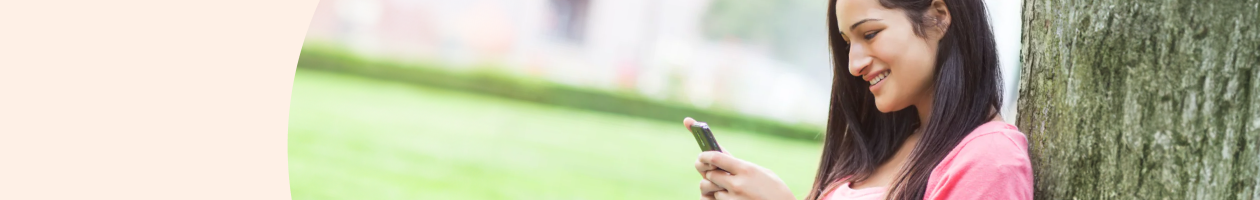 Young woman smiling and looking at her smartphone while leaning against a tree in a park.