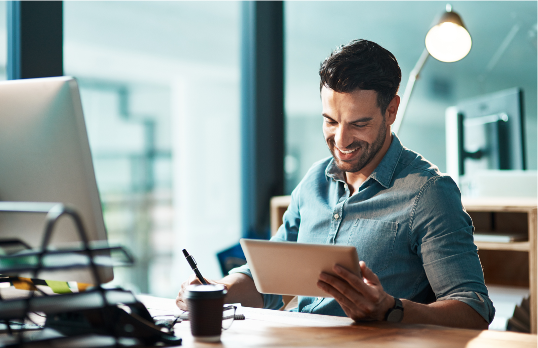 Man smiling while using a tablet at his desk with a computer, coffee cup, and desk lamp nearby.