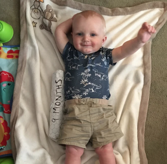 Baby lies on a blanket, wearing a blue dinosaur onesie and tan shorts, with a 9 months sign.