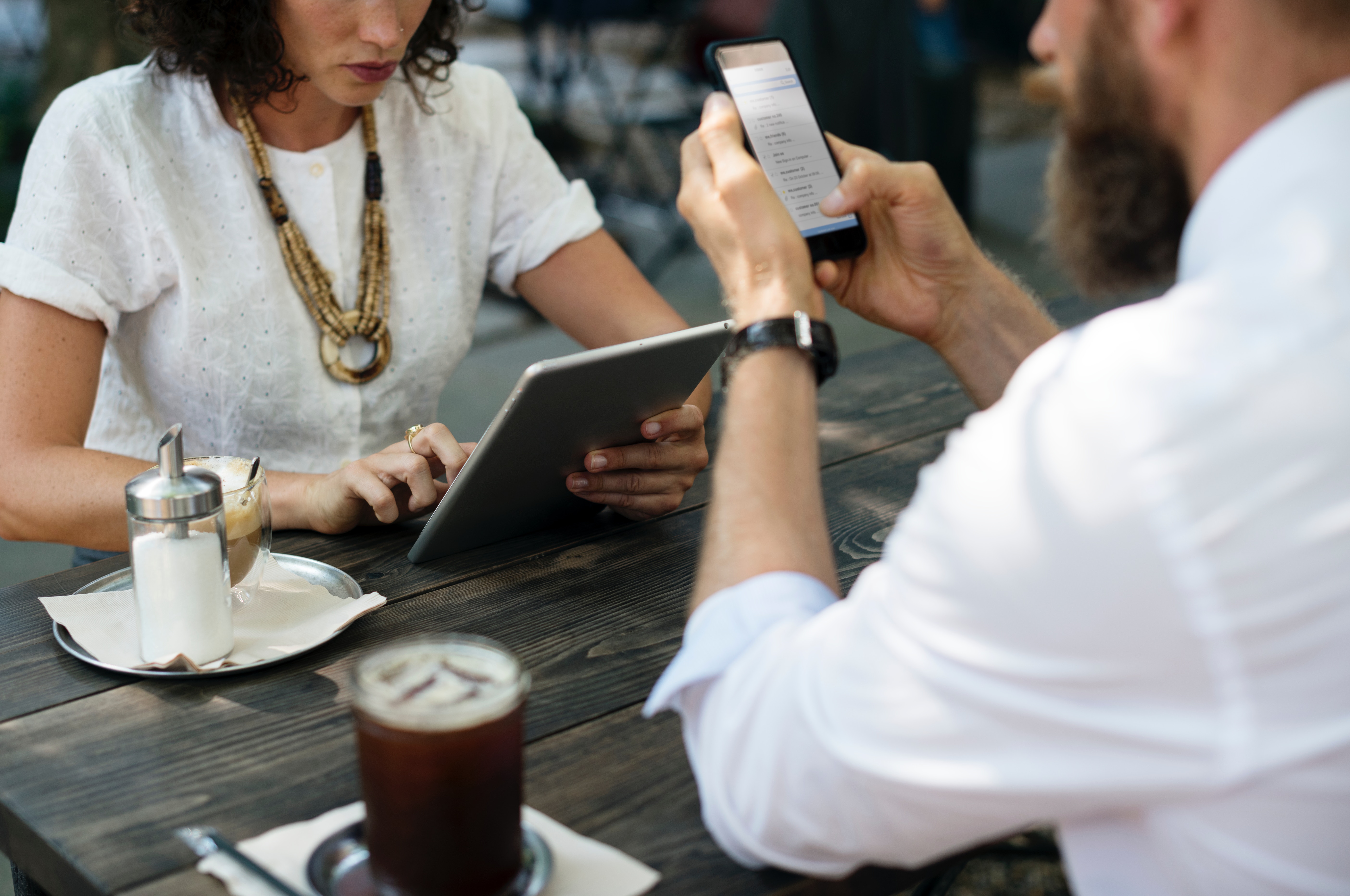 Two people using a tablet and smartphone at an outdoor cafe table with drinks and a sugar container.