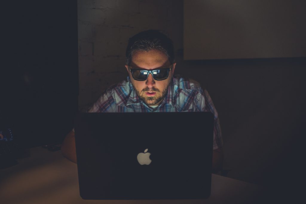 Man with sunglasses sitting in front of a laptop, illuminated by the screen in a dark room.