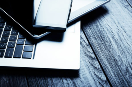 Close-up of a smartphone, tablet, and laptop stacked on a wooden desk.