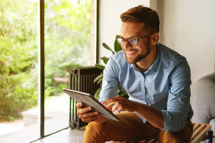 Smiling man with glasses using a tablet in a bright room near a large window.