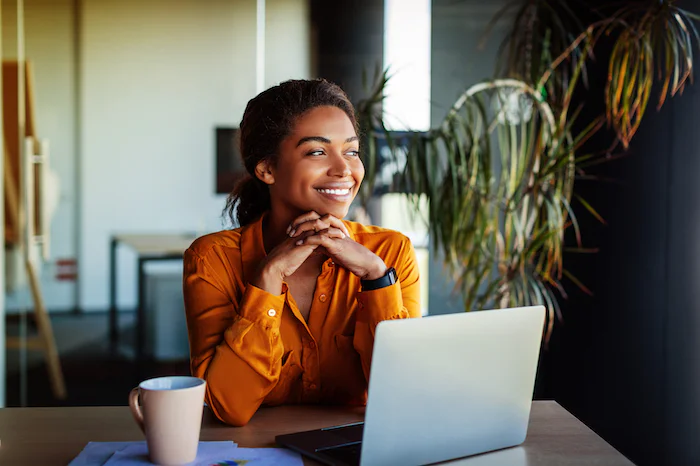 Smiling woman in orange shirt sitting at desk with laptop and cup, looking out window