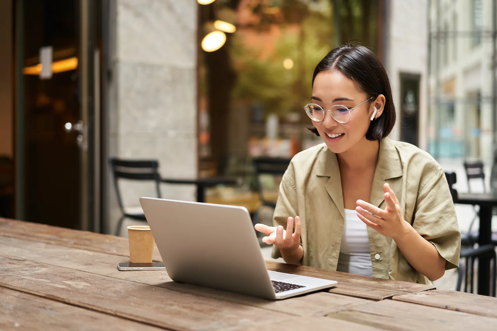 Young woman sitting on online meeting in outdoor cafe, talking to laptop camera, explaining something, drinking coffee. Digital nomad