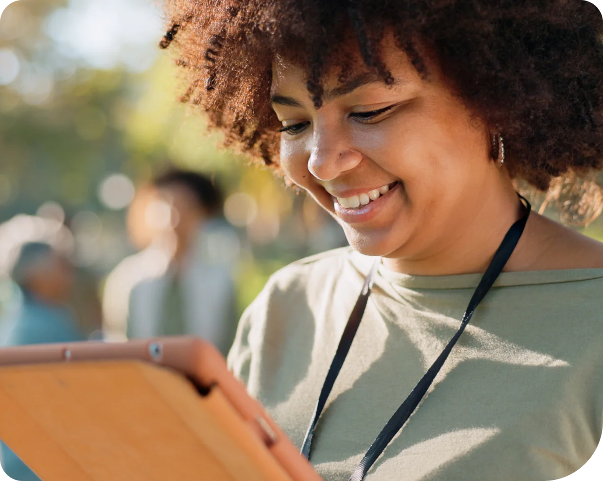 Smiling woman looking at tablet outdoors with trees in the background.