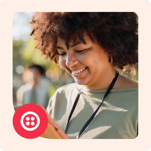 A woman with curly hair smiling at a tablet screen while standing outside with a blurred background.