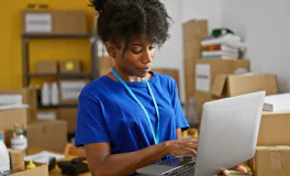 Woman with curly hair wearing a blue shirt and lanyard working on a laptop