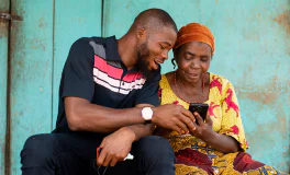 Young man smiling and showing elderly woman how to use her smartphone against a teal background.