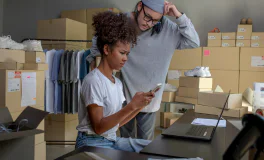 Two people working together in a stockroom with clothing and boxes, using a phone and a laptop.