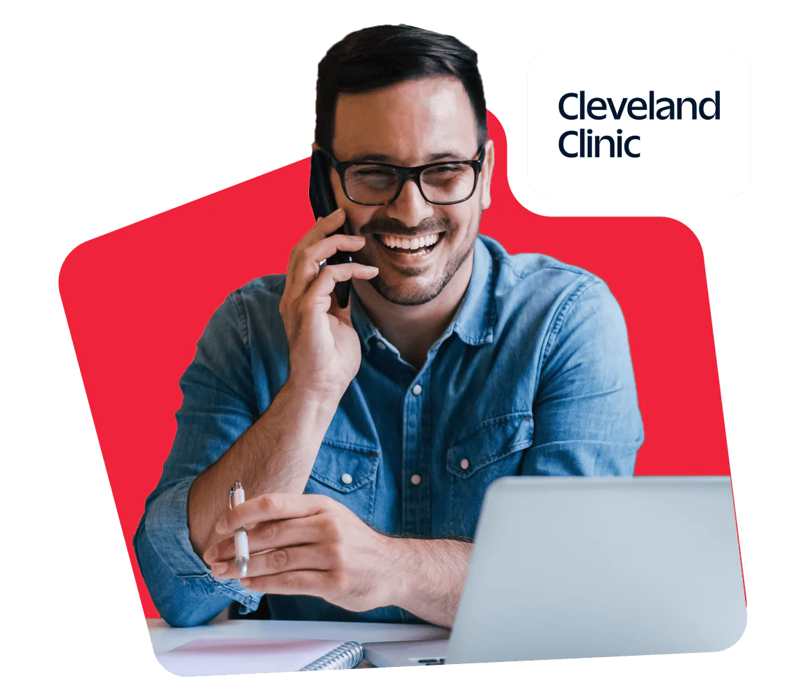 Man on phone working on laptop, Cleveland Clinic logo Smiling man talking on phone while using laptop with Cleveland Clinic logo in background.